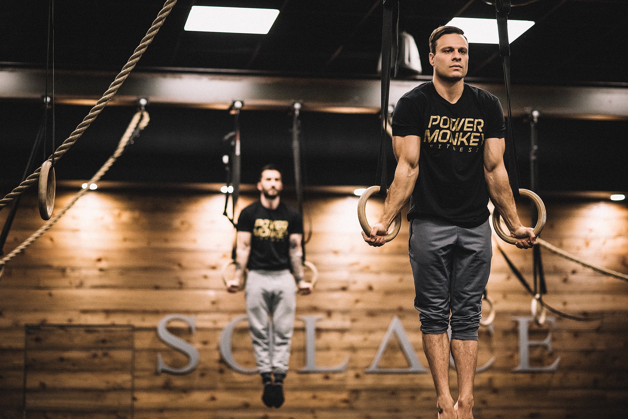 Photo of Dave Durante and Colin Geraghty performing a ring muscle up in a gym.
